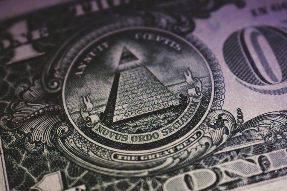 A close-up of the Great Seal on the back of a U.S. one-dollar bill, showing the pyramid with an eye at the top and the words 'Annuit Coeptis' and 'Novus Ordo Seclorum' in Latin, with detailed engraving patterns around the seal.