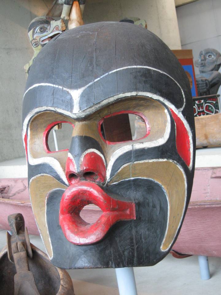A traditional Dzunukwa mask featuring bold black, red, and brown colors, with exaggerated facial features, including a round open mouth and large eyes, displayed in a museum setting.