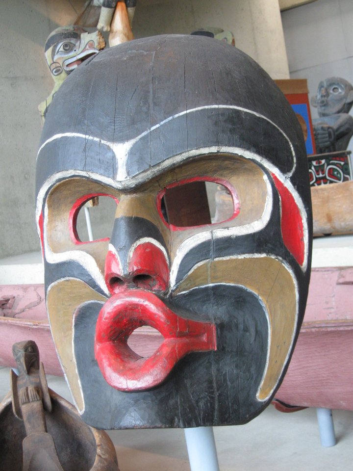 A traditional Dzunukwa mask featuring bold black, red, and brown colors, with exaggerated facial features, including a round open mouth and large eyes, displayed in a museum setting.