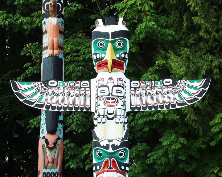 A colorful totem pole with intricate carvings of animals, including a large bird with outstretched wings at the top, painted in white, green, black, and red, standing against a backdrop of green trees.