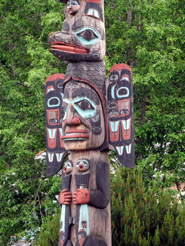 A colorful totem pole with intricate carvings of animals, including a large bird with outstretched wings at the top, painted in white, green, black, and red, standing against a backdrop of green trees.