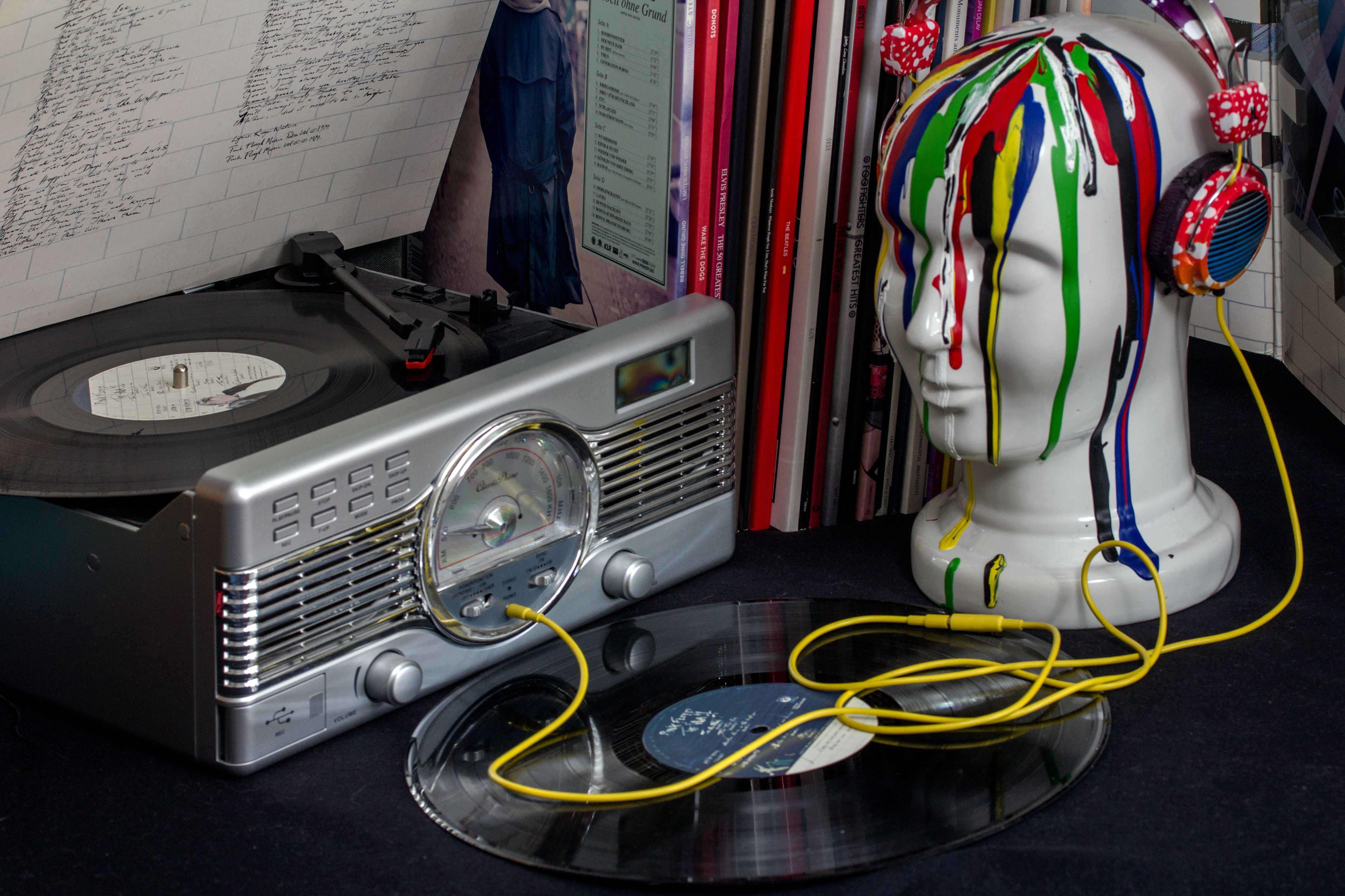 A vinyl record player spinning a record, with another vinyl placed in front of it. Beside the player is a white mannequin head with colorful paint drips and wearing red polka-dotted headphones connected to the record player by a yellow cable, against a backdrop of vinyl album covers.