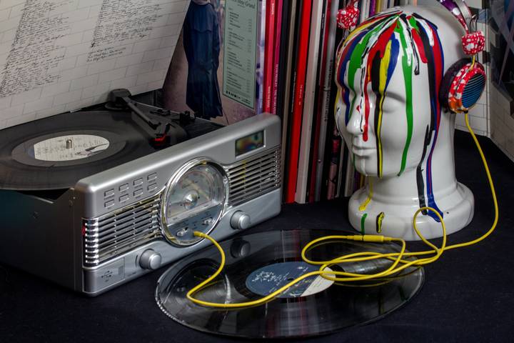 A vinyl record player spinning a record, with another vinyl placed in front of it. Beside the player is a white mannequin head with colorful paint drips and wearing red polka-dotted headphones connected to the record player by a yellow cable, against a backdrop of vinyl album covers.